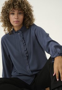 Blouse bleu marine avec un col froncé haut, fermeture à boutons sur le devant et poignets froncés. Tissu doux avec une texture légèrement brillante.