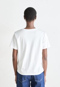 Tricou alb din bumbac, cu guler rotund și mâneci scurte, văzut din spate. Asortat cu blugi din denim albastru, ambele având un design simplu.