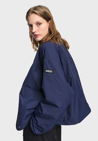 Jeune femme aux cheveux châtain clair portant une veste ample bleu marine avec un patch "AIGLE 1853" sur la manche, regardant par-dessus son épaule.