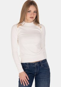 Giovane donna con lunghi capelli biondi indossa un maglione bianco aderente a maniche lunghe con bottoni dorati sulle spalle e jeans blu scuro.