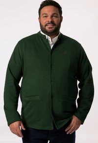 Mörkgrön kofta i mjukt tyg, med knappstängning fram, två framfickor och en diskret broderad logotyp på bröstet.
