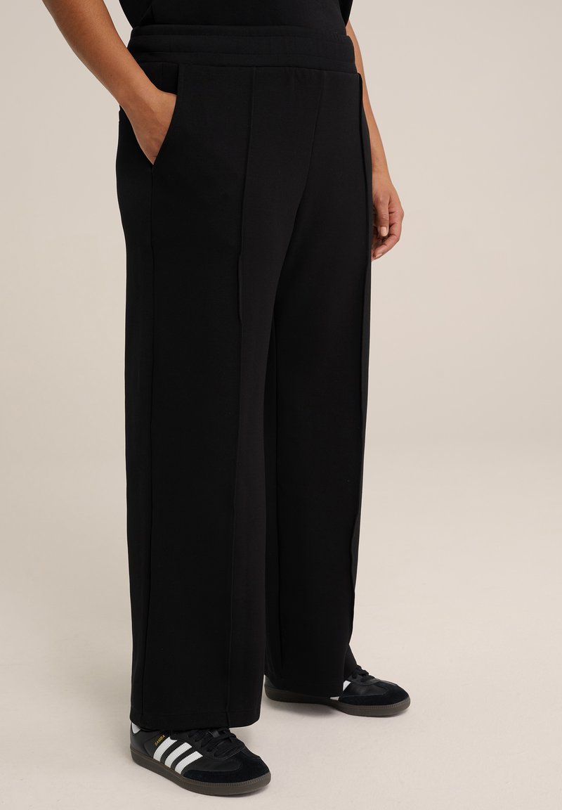 Pantalons larges noirs en tissu doux, dotés d'une taille haute, de poches latérales et d'une texture lisse. Assortis à des baskets noires.