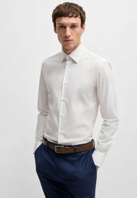 Camicia bianca a maniche lunghe con colletto classico, caratterizzata da una superficie liscia e chiusura a bottoni. Indossata con pantaloni blu navy e una cintura marrone.