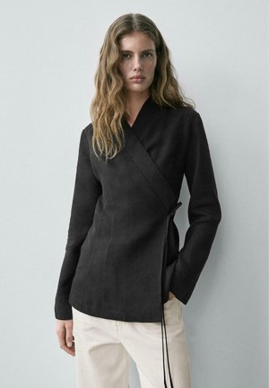 Donna con capelli mossi che indossa una giacca avvolgente aderente nera e pantaloni beige chiaro, in piedi davanti a uno sfondo grigio chiaro uniforme.