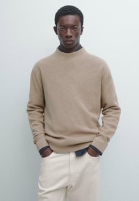 Beżowy prążkowany sweter z wysokim kołnierzem, długimi rękawami i ciemnoniebieskimi akcentami na mankietach, noszony na off-white spodniach na neutralnym tle.