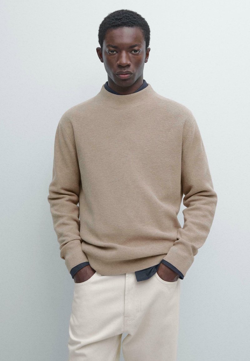 Beżowy prążkowany sweter z wysokim kołnierzem, długimi rękawami i ciemnoniebieskimi akcentami na mankietach, noszony na off-white spodniach na neutralnym tle.