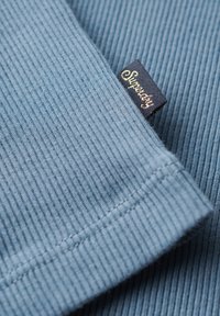 Blauwe ribbelstof met een zachte textuur. Bevat een zwart etiket met "Superdry" in gouden cursief, gelegen net bij de rand.