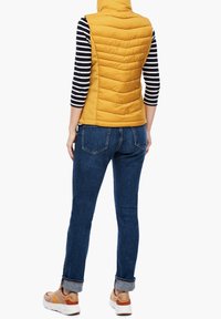 Żółta ocieplana kamizelka w pionowy pikowany wzór, noszona na czarno-białą koszulę z długim rękawem, zestawiona z ciemnoniebieskimi dżinsami i sneakerami.