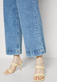 Jalkaterät, joissa on beigenväriset korolliset sandaalit, joissa on kaksi pehmeää hihnaa, yhdistettyinä leveisiin, lyhytlahkeisiin sinisiin farkkuihin tasaisella taustalla.