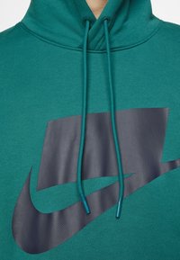 Teal huppari, jossa on suuri tummanharmaa logo. Varustettu säädettävillä nyöreillä ja teksturoidulla kankaalla mukavuutta varten.