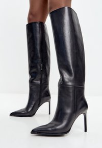 Bottes noires en cuir montantes avec un bout pointu, un design effilé et de hauts talons aiguilles. Comprend un détail de fermeture à glissière sur le côté.