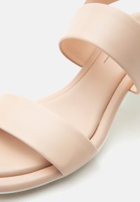 Κοντινό πλάνο μιας μπεζ σανδάλιας με δύο φαρδιές λωρίδες και ένα λείο, μαλακό πέλμα σε ανοιχτό φόντο.