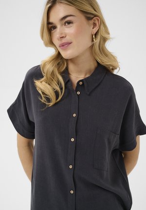 Šviesiaplaukė moteris su banguotais plaukais, vilkinti auksinius žiedinius auskarus ir tamsiai mėlyną trumpomis rankovėmis palaidinę su kišenėle krūtinėje, stovinti prieš baltą foną.