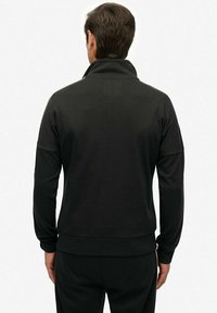 Sudadera negra con cremallera, cuello alto, mangas largas y dobladillo ajustado. Textura suave, diseño minimalista, sin patrones ni acentos visibles.
