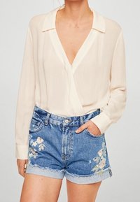 Blouse crème avec col, associée à un short en jean bleu orné de broderies florales et d'un ourlet effiloché. Texture de tissu lisse sur toute la surface.