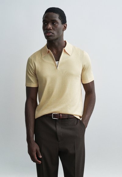 Homme portant un polo à manches courtes beige avec une fermeture éclair, un pantalon marron foncé et une ceinture tissée marron, posant devant un fond uni.