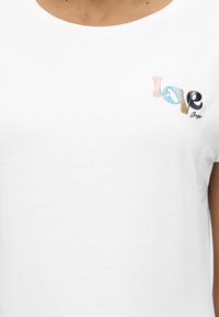 Λευκό T-shirt με πολύχρωμη κεντημένη λέξη "Love" στο αριστερό στήθος, φορεμένο από άτομο με ορατό λαιμό και άνω ώμους.