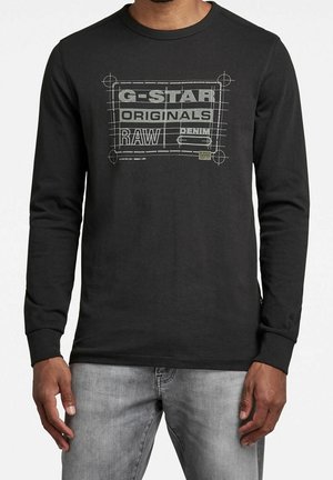 Mężczyzna w czarnej bluzie z długim rękawem z szarym nadrukiem „G-STAR ORIGINALS RAW DENIM” oraz jasnoszarymi dżinsami, stojący na jednolitym tle.