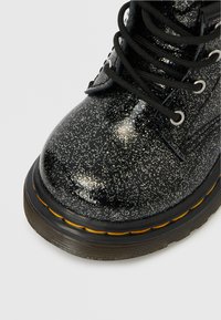 Μαύρο λαμπερό μποτάκι αστραγάλου με γυαλιστερό φινίρισμα, στρογγυλή μύτη, μαύρα κορδόνια, ασημένιες οπές και κίτρινη ραφή κατά μήκος της σόλας.