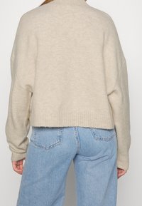 Personne portant un pull beige en peluche et un jean taille haute bleu clair, vu de dos contre un fond uni.