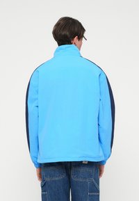 Ανοιχτό μπλε μπουφάν με ναυτικές λεπτομέρειες, ψηλό γιακά, ελαστικούς μανσέτες και χαλαρή εφαρμογή, κατασκευασμένο από απαλό, ελαφρύ υλικό.