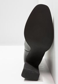 Chaussure à talon haut en cuir noir vue du dessous, présentant une surface lisse et une semelle en caoutchouc texturée avec des rainures horizontales.