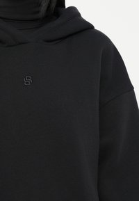 Sudadera negra con capucha hecha de tela suave, que presenta un logo bordado sutil en el pecho y un diseño de ajuste relajado.