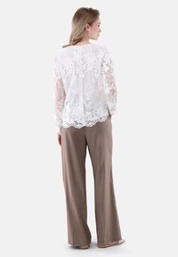 Weißes Spitzenoberteil mit floralen Stickereien, durchsichtigen Ärmeln und gewelltem Saum, kombiniert mit beigen, hochtaillierten, weiten Hosen.