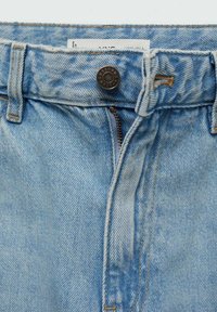 Gros plan sur la taille d'un jean en denim bleu clair avec le bouton fermé et la fermeture éclair partiellement ouverte.
