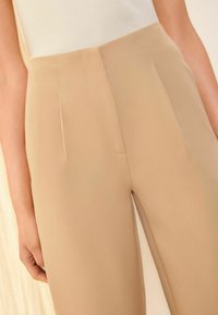 Beige tailored trousers made of smooth fabric feature front pleats and a straight leg design, with subtle stitching details near pockets.