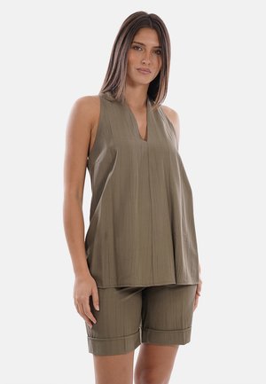 Donna che indossa un top verde oliva senza maniche e pantaloncini coordinati di lunghezza fino al ginocchio su uno sfondo bianco semplice.