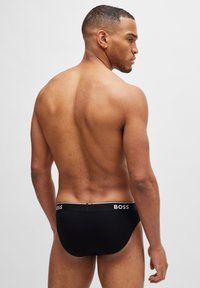 Schwarze Slip-Unterwäsche mit einem Logo-Bund, gestaltet mit einer glatten Textur und einer figurschmeichelnden Passform, von hinten präsentiert.