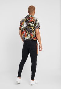 Camisa de manga corta con un patrón abstracto multicolor que incluye negro, rojo, amarillo y verde. Combinada con jeans slim-fit negros y zapatillas blancas.