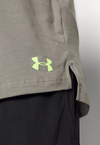 Detail šedého trička Under Armour se zapínáním a neonovým logem na černých texturovaných sportovních šortkách.