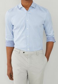 Chemise boutonnée à rayures bleu clair et blanc avec manches retroussées, associée à un pantalon gris clair. Tissu en coton avec une texture lisse.