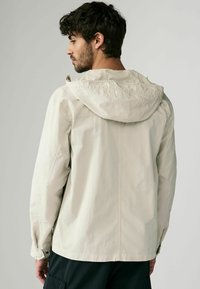 Jasno beżowa wodoodporna kurtka z kapturem, długimi rękawami i elastycznymi mankietami. Gładka faktura i minimalistyczny design bez widocznych wzorów.