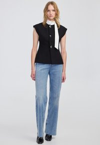 Czarny bezrękawnik z guzikami z przodu i białym krawatem na szyi, w połączeniu z jasnoniebieskimi dżinsami w szerszym kroju i czarnymi botkami za kostkę.