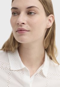Chemise en coton blanc avec détails brodés et découpés, col à boutons et texture lisse, en gros plan sur le visage et le cou d'un mannequin.