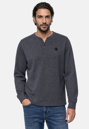 Homme aux cheveux foncés et barbe, portant un t-shirt henley gris foncé à manches longues et un jean bleu, debout, une main dans la poche.