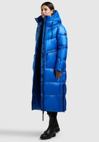 Mėlynas pūkinis paltas su ilgu, per didelio dydžio dizainu. Turi aukštą apykaklę, kišenes su užtrauktukais ir blizgią išorinę dalį. Dėvimas su juodais kulkšnies batais.