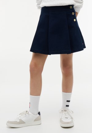 Marineblå plisseret nederdel med guldknapper på siden, båret med hvide sneakers og hvide crew-sokker med sorte logo-striber.