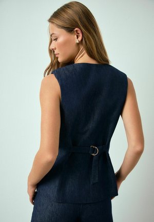 Kobieta ubrana w ciemny jeansowy top bez rękawów z materiałowym paskiem i metalową sprzączką, pokazana od tyłu na jasnym, jednolitym tle.