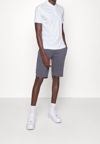 Vit bomullspikétröja med knäppkrage, kombinerad med gråa shorts. Vita sneakers och fotlångstrumpor fulländar outfiten.