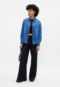 Camisa de ganga com botões de pressão, dois bolsos no peito e mangas longas, combinada com calças largas pretas e uma pequena mala brilhante.