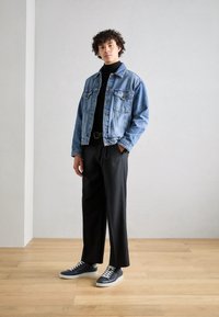 Veste en denim, bleu clair, quatre poches, associée à un col roulé noir et un large pantalon noir. Baskets grises avec des lacets blancs. Fond neutre.