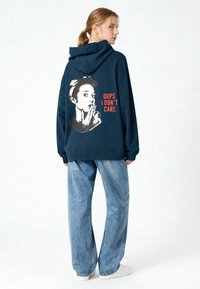 Felpa oversize blu navy realizzata in tessuto morbido, con una grafica di una donna e il testo "OOPS I DON'T CARE" in arancione sul retro.