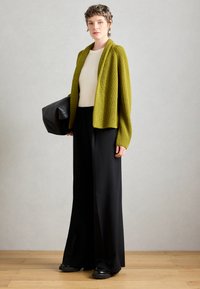 Cardigan a coste verde oliva sopra una maglietta crema, abbinato a pantaloni neri a gamba larga e scarpe nere chunky, mentre tiene una borsa nera.