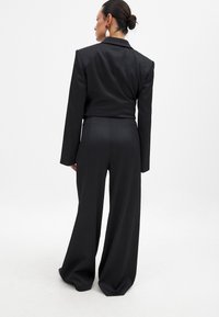 Črn kakovostno krojen suit z skrajšanim jopičem in širokimi hlačami. Gladek material, zarezan ovratnik in dolgi rokavi. Minimalistična zasnova.