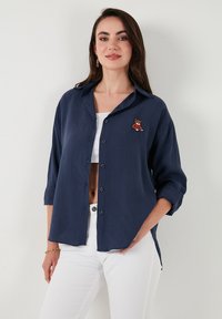 Navy button-up shirt with rolled sleeves and an embroidered bear detail. Made of lightweight fabric, featuring a relaxed fit and curved hem.
