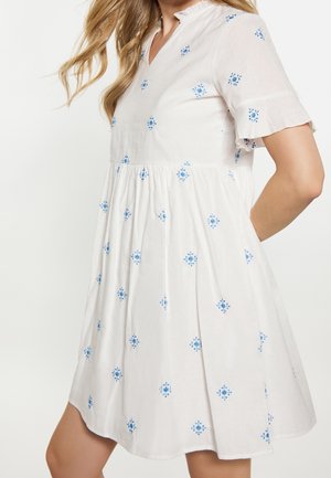 Witte jurk tot op de knie met korte geruwde mouwen en blauwe bloemrijke borduursels, gedragen door een persoon met lang blond haar.